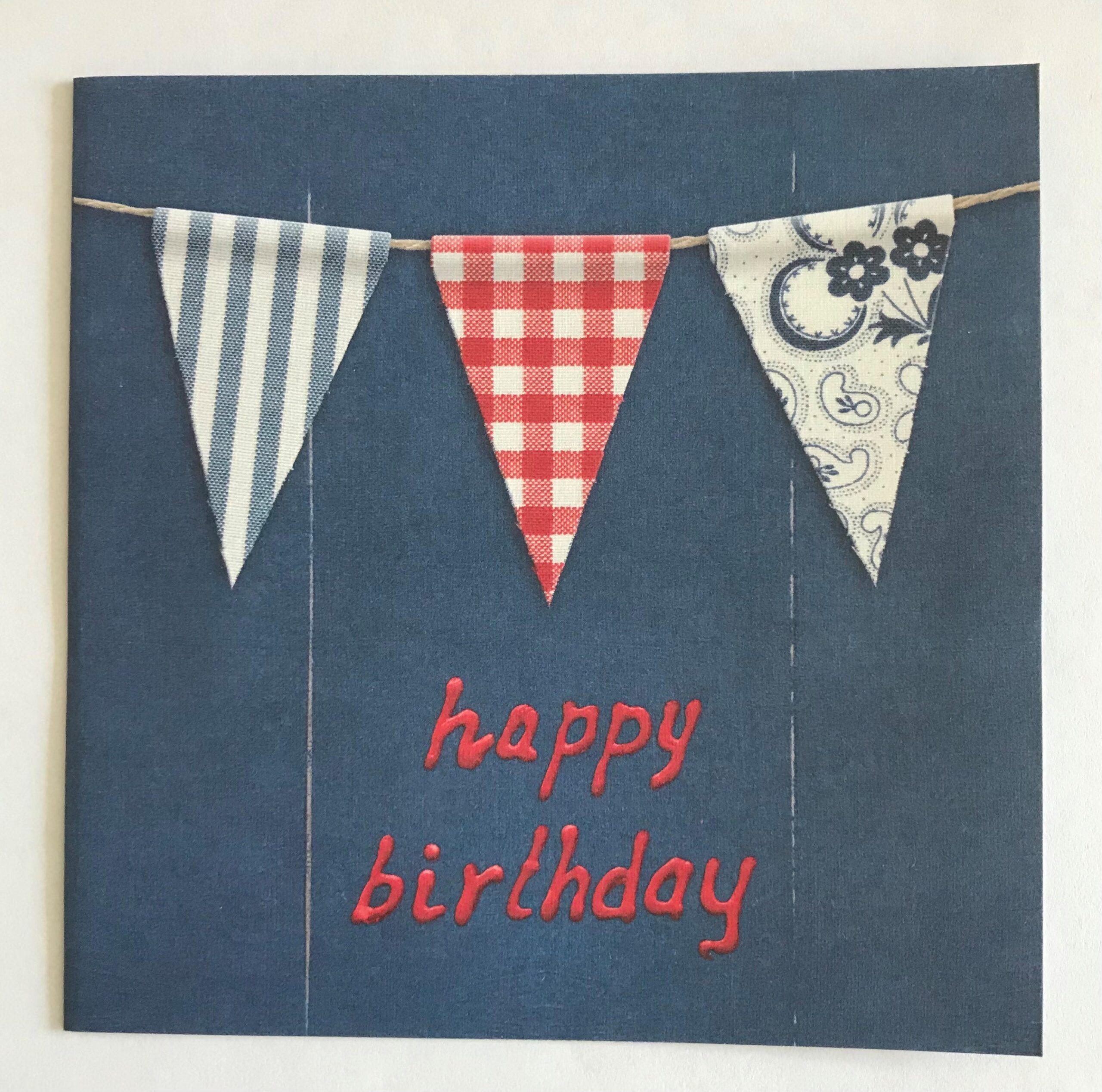 Op een blauwe kaart staat in rode letters "Happy birthday". Daarbij is een slinger afgebeeld met drie driehoekige vlaggetjes eraan: een blauw/wit gestreepte, een rood/wit geblokte en een met bloemen in wit met blauw. De envelop is rood/wit geblokt.